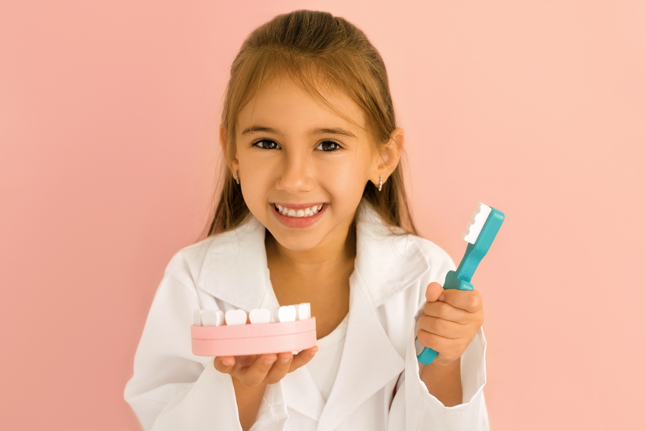 A little dentist girl holds a model of a human jaw and a toothbrush in her hands to demonstrate proper tooth brushing isolated on pink studio background. International Dentist's Day